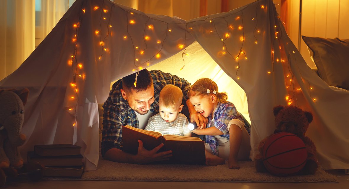 Erwachsene Person mit zwei Kindern in beleuchtetem Zelt aus Buch vorlesend